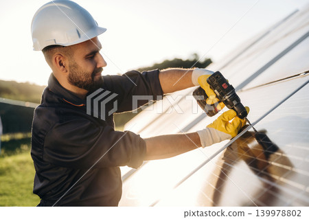Yellow drill is in hands, installation process. Male solar engineer examining photovoltaic panels at a power station Yellow drill is in hands, installation process. Male solar engineer examining photovoltaic panels at a power station 139978802