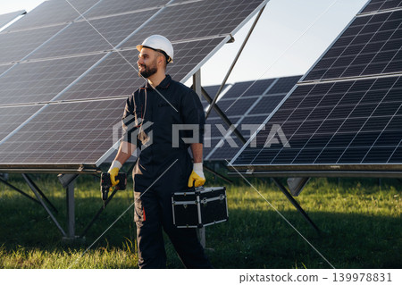 Box in hand, walking. Male solar engineer examining photovoltaic panels at a power station Box in hand, walking. Male solar engineer examining photovoltaic panels at a power station 139978831