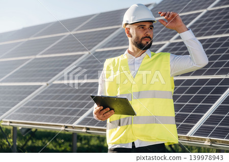 In yellow colored professional uniform. Male solar engineer examining photovoltaic panels at a power station In yellow colored professional uniform. Male solar engineer examining photovoltaic panels at a power station 139978943