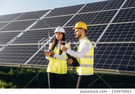 Windmill little model is in hands of woman. Two solar engineers standing together at a photovoltaic field, in bright safety vests Windmill little model is in hands of woman. Two solar engineers standing together at a photovoltaic field, in bright safety vests 139979061