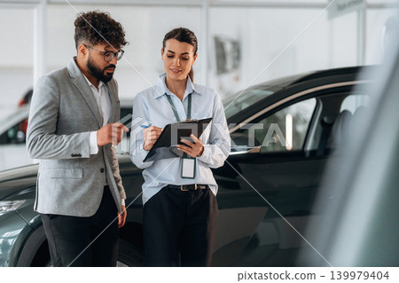 Holding notepad with information. Business consultation between car dealer and buyer in showroom Holding notepad with information. Business consultation between car dealer and buyer in showroom 139979404
