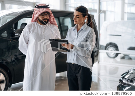 At daytime, showing information from notepad. Middle eastern client discussing car purchase in modern dealership At daytime, showing information from notepad. Middle eastern client discussing car purchase in modern dealership 139979500