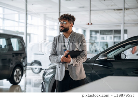 Standing, front view. Confident car salesman using digital tablet inside modern showroom Standing, front view. Confident car salesman using digital tablet inside modern showroom 139979555