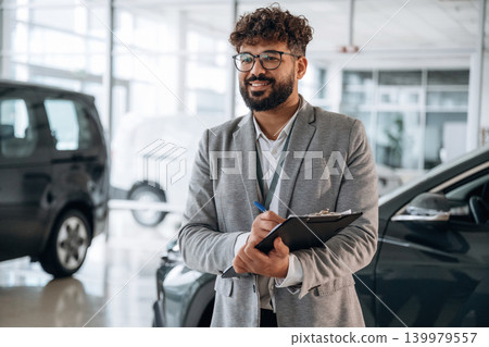 Standing, front view. Confident car salesman using digital tablet inside modern showroom Standing, front view. Confident car salesman using digital tablet inside modern showroom 139979557