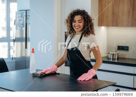 In uniform. White woman cleaning the kitchen countertop with spray and cloth wearing pink gloves In uniform. White woman cleaning the kitchen countertop with spray and cloth wearing pink gloves 139979669