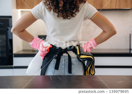 Woman wearing cleaning gloves and apron standing in a modern kitchen, preparing for household chores 139979695