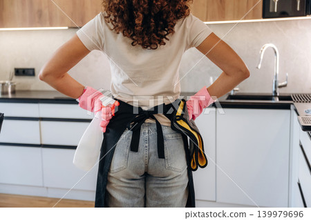 Woman wearing cleaning gloves and apron standing in a modern kitchen, preparing for household chores 139979696