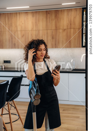 Woman wearing an apron and cleaning gloves taking a break and using her smartphone in a kitchen Woman wearing an apron and cleaning gloves taking a break and using her smartphone in a kitchen 139979703