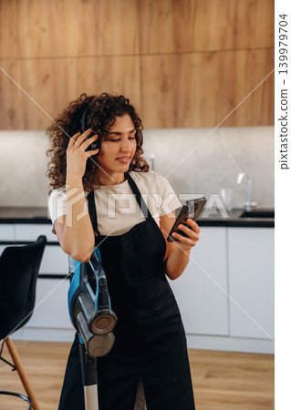 Woman wearing an apron and cleaning gloves taking a break and using her smartphone in a kitchen Woman wearing an apron and cleaning gloves taking a break and using her smartphone in a kitchen 139979704