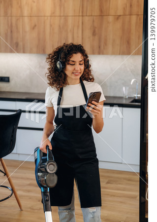 Woman wearing an apron and cleaning gloves taking a break and using her smartphone in a kitchen Woman wearing an apron and cleaning gloves taking a break and using her smartphone in a kitchen 139979705
