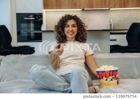 Weekend activities. Happy woman relaxing on a sofa with popcorn, watching TV and enjoying her leisure time at home Weekend activities. Happy woman relaxing on a sofa with popcorn, watching TV and enjoying her leisure time at home 139979754