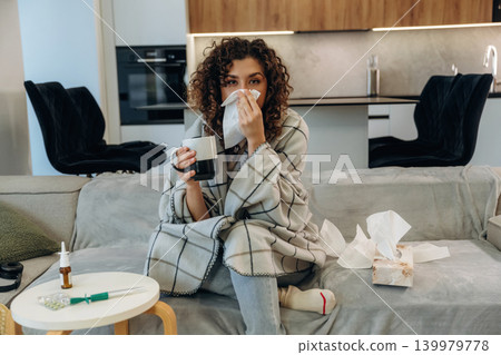 White cup is in hand. Sick woman feeling unwell at home, checking her temperature and using medicine while resting on a sofa White cup is in hand. Sick woman feeling unwell at home, checking her temperature and using medicine while resting on a sofa 139979778