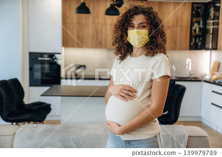 In a mask. A pregnant woman at home, holding her belly and enjoying a peaceful moment in a modern kitchen In a mask. A pregnant woman at home, holding her belly and enjoying a peaceful moment in a modern kitchen 139979789