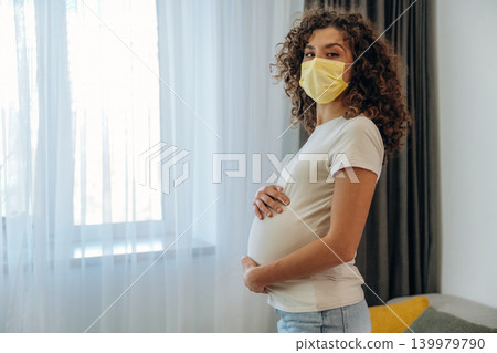 In a mask. A pregnant woman at home, holding her belly and enjoying a peaceful moment in a modern kitchen 139979790