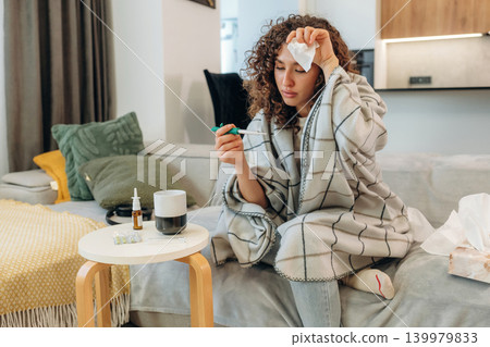 Sick woman feeling unwell at home, checking her temperature and using medicine while resting on a sofa Sick woman feeling unwell at home, checking her temperature and using medicine while resting on a sofa 139979833