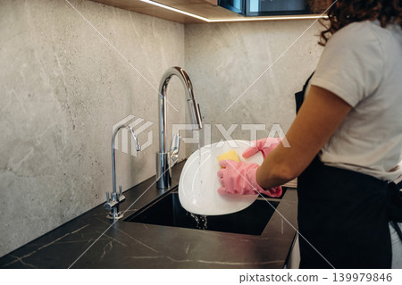Pink colored gloves. In domestic room. Woman washing dishes with sponge in modern kitchen Pink colored gloves. In domestic room. Woman washing dishes with sponge in modern kitchen 139979846