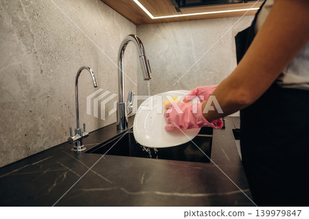 Pink colored gloves. In domestic room. Woman washing dishes with sponge in modern kitchen Pink colored gloves. In domestic room. Woman washing dishes with sponge in modern kitchen 139979847