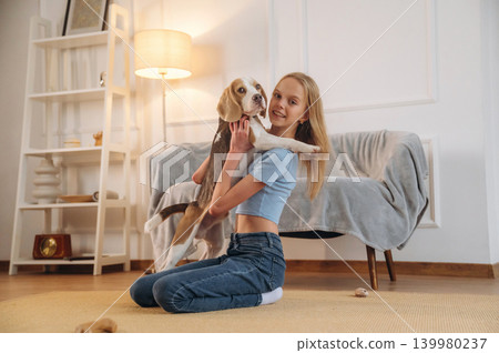 Side view, sitting. Girl is with her dog on the floor in a cozy living room Side view, sitting. Girl is with her dog on the floor in a cozy living room 139980237
