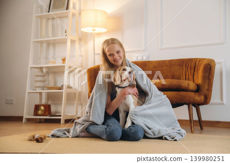 Covering themselves with a blanket. Girl is with her dog on the floor in a cozy living room Covering themselves with a blanket. Girl is with her dog on the floor in a cozy living room 139980251