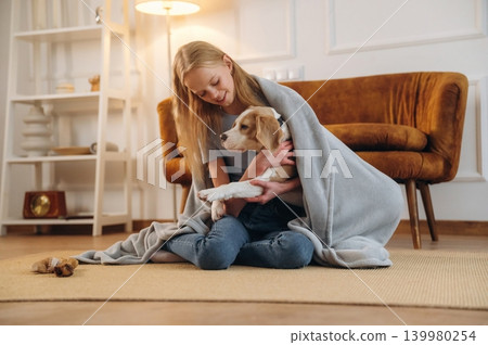 Covering themselves with a blanket. Girl is with her dog on the floor in a cozy living room Covering themselves with a blanket. Girl is with her dog on the floor in a cozy living room 139980254