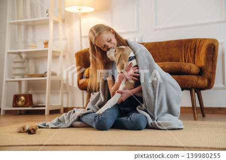 Covering themselves with a blanket. Girl is with her dog on the floor in a cozy living room Covering themselves with a blanket. Girl is with her dog on the floor in a cozy living room 139980255