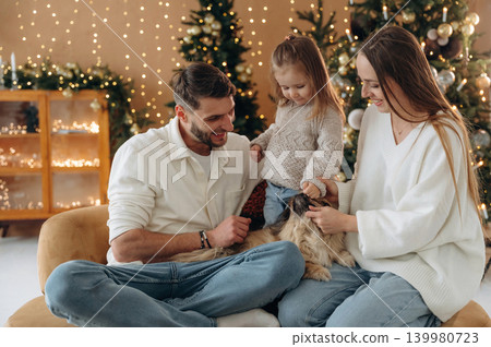Pet is on the legs. Happy family with child and dog celebrating Christmas together at home with decorated tree 139980723