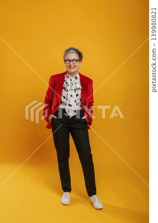 Confident woman with stylish gray hair wearing a red blazer and polka dot blouse stands against a vibrant yellow background, showcasing modern fashion and self-expression Confident woman with stylish gray hair wearing a red blazer and polka dot blouse stands against a vibrant yellow background, showcasing modern fashion and self-expression 139980821