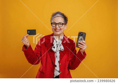 Senior woman in stylish red blazer holding smartphone and credit card, smiling against vibrant yellow background, showcasing modern financial technology and empowerment Senior woman in stylish red blazer holding smartphone and credit card, smiling against vibrant yellow background, showcasing modern financial technology and empowerment 139980826