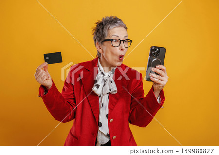 Excited older woman in stylish red blazer holds smartphone and credit card, expressing joy and surprise against vibrant yellow background, showcasing modern technology use Excited older woman in stylish red blazer holds smartphone and credit card, expressing joy and surprise against vibrant yellow background, showcasing modern technology use 139980827