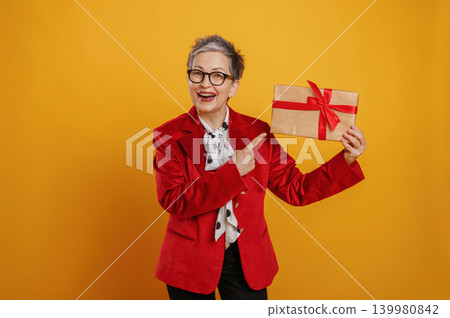 Senior woman in red blazer joyfully holding a gift box with a red ribbon against a vibrant yellow background, celebrating the spirit of giving and joy Senior woman in red blazer joyfully holding a gift box with a red ribbon against a vibrant yellow background, celebrating the spirit of giving and joy 139980842