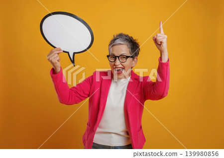 Excited woman in pink blazer holds speech bubble prop, celebrating with raised finger against vibrant yellow background, conveying enthusiasm and creativity 139980856