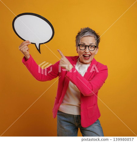 Senior woman with short gray hair is joyfully holding a speech bubble prop against a vibrant yellow background, expressing excitement and creativity in a playful manner 139980857