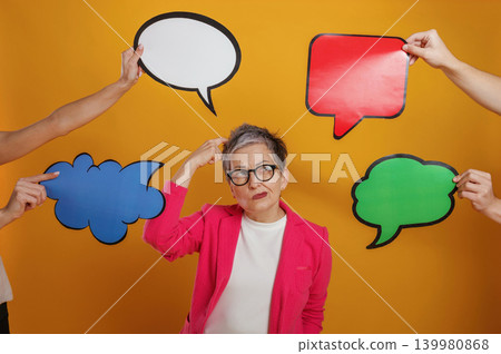 Senior woman in pink blazer looks confused while surrounded by colorful speech bubbles, representing communication challenges and decision-making in a vibrant setting Senior woman in pink blazer looks confused while surrounded by colorful speech bubbles, representing communication challenges and decision-making in a vibrant setting 139980868