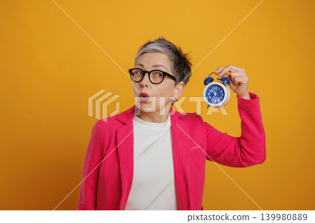 Senior woman in vibrant pink blazer holds blue alarm clock, expressing surprise against a bright yellow background, showcasing the concept of time management and urgency Senior woman in vibrant pink blazer holds blue alarm clock, expressing surprise against a bright yellow background, showcasing the concept of time management and urgency 139980889