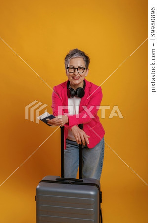 Senior woman with stylish glasses and headphones is smiling while holding a passport and standing next to a suitcase against a vibrant yellow background Senior woman with stylish glasses and headphones is smiling while holding a passport and standing next to a suitcase against a vibrant yellow background 139980896