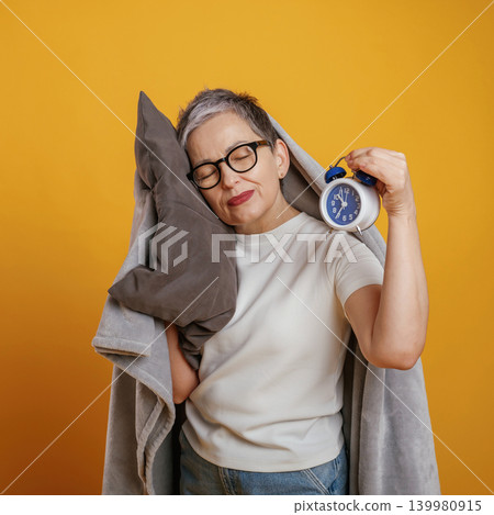 Mature woman with short gray hair embraces a pillow while holding an alarm clock, conveying a sense of relaxation and comfort against a vibrant yellow backdrop Mature woman with short gray hair embraces a pillow while holding an alarm clock, conveying a sense of relaxation and comfort against a vibrant yellow backdrop 139980915