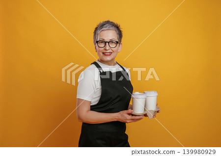 Smiling woman with short gray hair wearing black apron holds coffee cups against vibrant yellow background, showcasing barista skills and customer service Smiling woman with short gray hair wearing black apron holds coffee cups against vibrant yellow background, showcasing barista skills and customer service 139980929