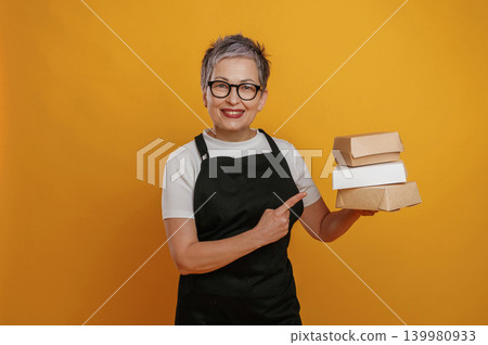 Smiling woman with short gray hair wearing glasses holds stacked takeout boxes against a vibrant yellow background, showcasing food packaging and delivery options Smiling woman with short gray hair wearing glasses holds stacked takeout boxes against a vibrant yellow background, showcasing food packaging and delivery options 139980933