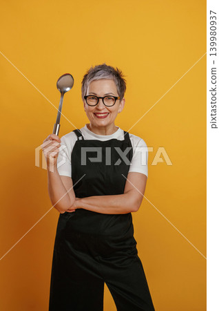 Cheerful woman chef with short gray hair is confidently holding a cooking spoon while wearing a black apron against a vibrant yellow background, showcasing her culinary passion and creativity 139980937