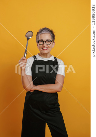 Confident woman chef with short gray hair holding a cooking spoon, wearing black apron against vibrant yellow background, showcasing culinary passion and creativity 139980938