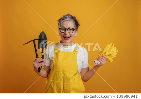 Senior woman gardener in bright yellow overalls smiles while holding gardening tools and gloves against a vibrant yellow background, showcasing gardening enthusiasm Senior woman gardener in bright yellow overalls smiles while holding gardening tools and gloves against a vibrant yellow background, showcasing gardening enthusiasm 139980941
