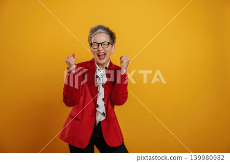 Joyful senior woman in red blazer celebrates success with raised fists against a vibrant yellow background, expressing happiness and excitement in a lively atmosphere 139980982