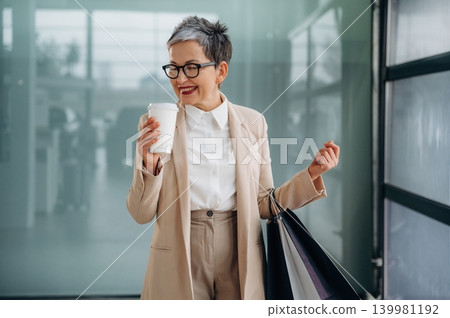Stylish older woman in beige suit holding coffee cup and shopping bags, smiling joyfully in a modern urban environment with glass walls and bright lighting Stylish older woman in beige suit holding coffee cup and shopping bags, smiling joyfully in a modern urban environment with glass walls and bright lighting 139981192
