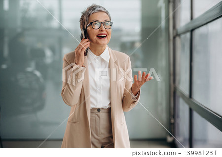 Senior businesswoman in stylish beige suit is joyfully talking on smartphone while standing in modern office space, showcasing professional communication and confidence Senior businesswoman in stylish beige suit is joyfully talking on smartphone while standing in modern office space, showcasing professional communication and confidence 139981231