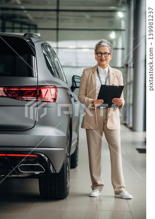 Senior woman in stylish suit stands confidently beside luxury SUV, holding clipboard in modern automotive showroom, showcasing professionalism and expertise in the automotive industry Senior woman in stylish suit stands confidently beside luxury SUV, holding clipboard in modern automotive showroom, showcasing professionalism and expertise in the automotive industry 139981237