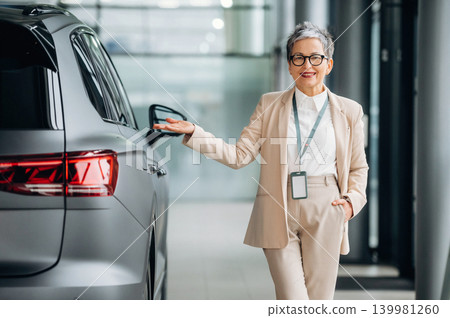 Confident senior woman in business attire presenting a sleek modern vehicle in a contemporary showroom, showcasing automotive sales and customer service experience Confident senior woman in business attire presenting a sleek modern vehicle in a contemporary showroom, showcasing automotive sales and customer service experience 139981260