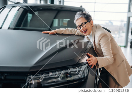 Senior woman in stylish attire joyfully embracing modern car, showcasing excitement and connection with automotive innovation in a bright showroom 139981286