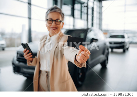 Senior woman in stylish outfit is smiling and holding a credit card in a modern car dealership, showcasing a confident purchasing experience with vehicles in background Senior woman in stylish outfit is smiling and holding a credit card in a modern car dealership, showcasing a confident purchasing experience with vehicles in background 139981305