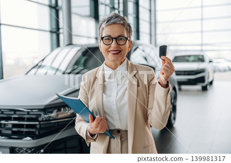 Confident senior woman in business attire is holding car key and clipboard, smiling in modern car dealership with vehicles in the background, showcasing customer service Confident senior woman in business attire is holding car key and clipboard, smiling in modern car dealership with vehicles in the background, showcasing customer service 139981317