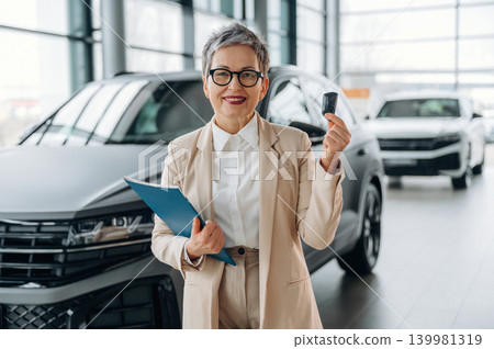 Confident senior woman in stylish suit holding car keys and clipboard, standing in modern showroom with luxury vehicles, representing automotive sales and customer service Confident senior woman in stylish suit holding car keys and clipboard, standing in modern showroom with luxury vehicles, representing automotive sales and customer service 139981319